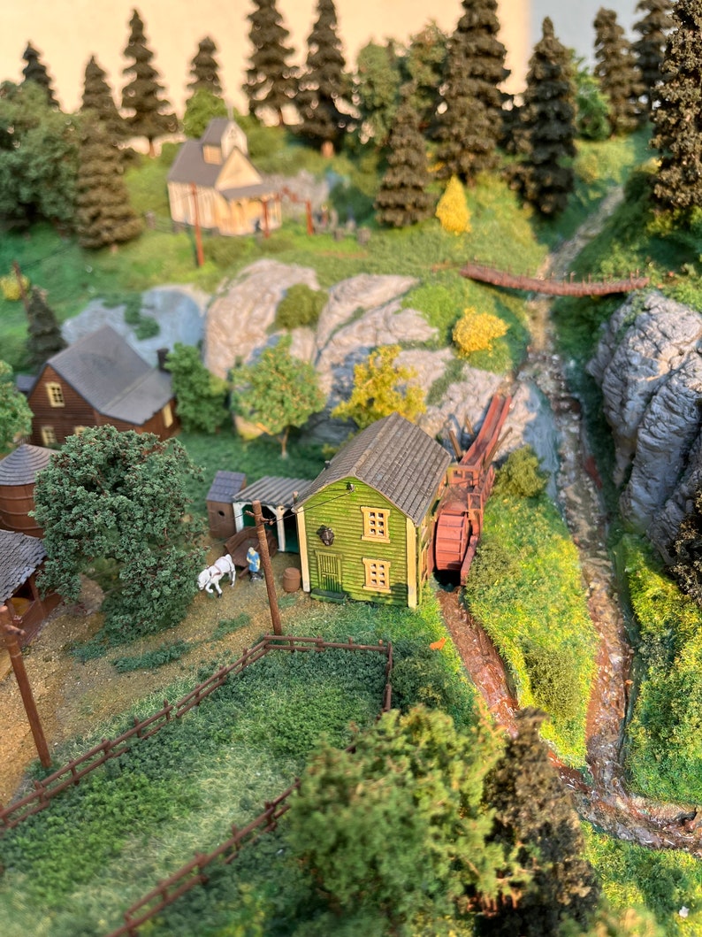 Pu&ograve; includere: Un modello in miniatura di una piccola citt&agrave; con un mulino ad acqua in legno verde, un ponte in legno e un ruscello che attraversa la citt&agrave;. Il modello &egrave; situato in una zona boscosa con alberi e rocce.