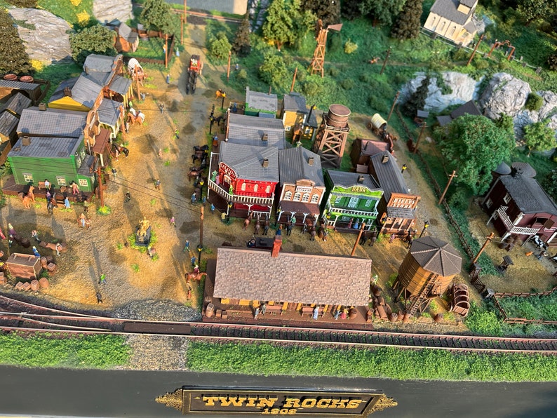 Pu&ograve; includere: Un modello in miniatura di una citt&agrave; western chiamata "Twin Rocks 1808" con edifici in legno, una torre d'acqua e un binario ferroviario. La citt&agrave; &egrave; circondata da alberi e rocce.