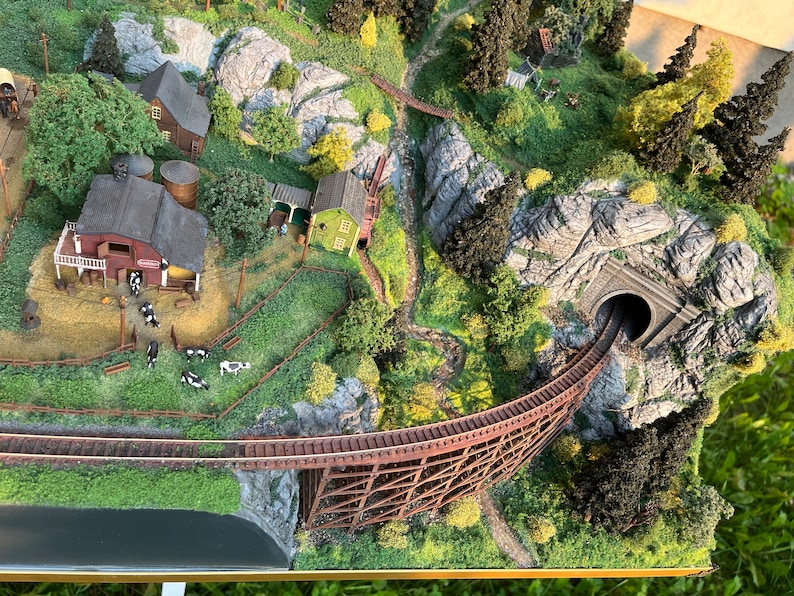 Pu&ograve; includere: Un set di treni in miniatura con un ponte in legno a traliccio, una galleria e una scena di fattoria con edifici e bestiame. La scena &egrave; ambientata in una zona montuosa con rocce e alberi.