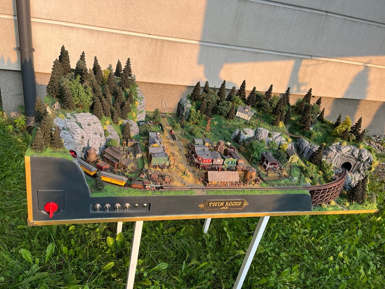 Pu&ograve; includere: Un set di treni in miniatura con una citt&agrave;, montagne e un tunnel. Il treno &egrave; giallo e nero e la citt&agrave; &egrave; composta da edifici in miniatura. Il set &egrave; su una base di legno con un cartello che dice "Twin Rocks Line".