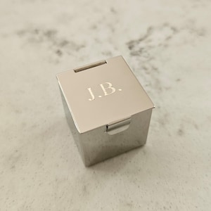 May include: A silver ring box with a hinged lid. The lid is engraved with the initials "J.B."
