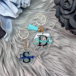 Op de afbeelding: Twee gepersonaliseerde sleutelhangers met goudkleurige hardware. Eén sleutelhanger heeft de naam "Savion" in blauw schrift op een grijze achtergrond, en de andere zegt "Danielle" in turquoise schrift. Beide sleutelhangers hebben een heldere, zeshoekige vorm.
