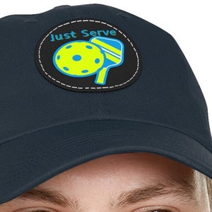 Può includere: Cappellino da baseball blu navy con una toppa circolare con un design di pickleball e racchetta gialli, con le parole "Just Serve" in bianco. La toppa è delineata da cuciture bianche e nere.