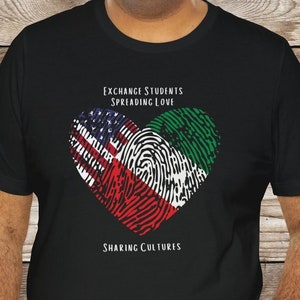 Puede incluir: Camiseta negra con un diseño en forma de corazón que presenta las banderas de Estados Unidos y Nigeria. El texto "Exchange Students Spreading Love" y "Sharing Cultures" está impreso en la camiseta.