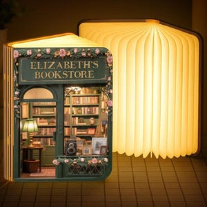 May include: A book-shaped lamp that opens to reveal a miniature bookstore interior. The dark green facade features the text ELIZABETH'S BOOKSTORE, pink floral accents, and a detailed interior with shelves, a lamp, and a camera. The pages emit a warm glow.