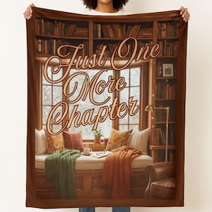 May include: A cozy throw blanket featuring a library scene with a window seat, bookshelves, and the text "Just One More Chapter" in elegant script. The blanket includes pillows, a cup, and a blanket in shades of orange, green, and red.