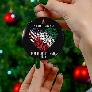Könnte beinhalten: Ein runder Weihnachtsbaumschmuck mit schwarzem Hintergrund und einem herzförmigen Design. Das Herz besteht aus zwei Fingerabdrücken, einer mit der amerikanischen Flagge und der andere mit der Flagge Saudi-Arabiens. Der Text "In every exchange love leaves its mark 2023" ist unter dem Herzen gedruckt.