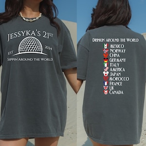 Op de afbeelding: Een donkergrijs T-shirt met een witte afbeelding van een golfbal en de tekst "Jessyka's 21st Est 2024 Sippin' Around the World". De achterkant van het shirt heeft de tekst "Drinkin' Around the World" met een lijst van landen en hun vlaggen.