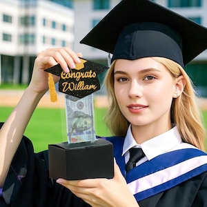 May include: A black graduation cap with gold glitter and the text "Congrats William" is attached to a clear plastic box with a dollar bill inside. A person wearing a black graduation gown and cap is holding the box.
