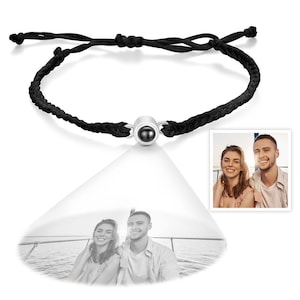 May include: Black braided cord bracelet with a silver-colored central bead. The bead appears to project an image of a couple. A small photo of the couple is in the corner. The bracelet is on a white background.