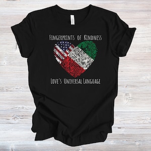 May include: Black t-shirt with a white heart-shaped fingerprint design. The fingerprint is made up of the American and Nigerian flags. The text "Fingerprints of Kindness" and "Love's Universal Language" is printed in white on the shirt.