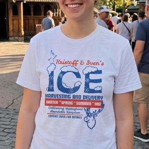 Könnte beinhalten: Ein weißes Kurzarm-T-Shirt mit einer blau-roten Grafik mit der Aufschrift Kristoff & Sven's ICE HARVESTING AND DELIVERY. Das Design zeigt eine kleine Olaf-Figur, das Wort ICE mit Schneeeffekten, saisonale Begriffe und eine Zeichnung von Sven.