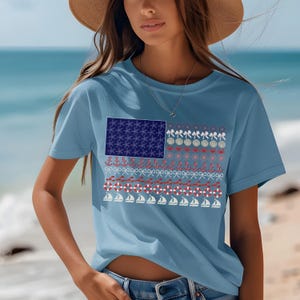 May include: Light blue t-shirt featuring a nautical-themed American flag design. The flag's stars are replaced with blue starfish, and stripes with anchors, bows, life preservers, and sailboats. The shirt is a short-sleeved crew neck.