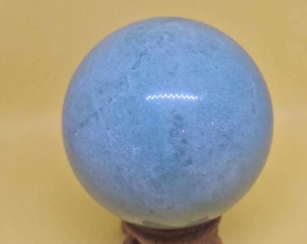 Puede incluir: Una esfera de cristal azul claro sobre un soporte de madera. La esfera es lisa y pulida, con un ligero brillo.