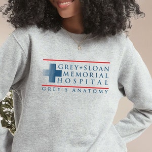 May include: A light gray sweatshirt featuring the text "GREY + SLOAN MEMORIAL HOSPITAL GREY'S ANATOMY" in blue and red. The sweatshirt has long sleeves and a crew neck.