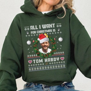 Peut inclure: Sweat-shirt à capuche vert avec un motif de Noël représentant un visage d'homme dans une couronne, des flocons de neige et le texte "All I want for Christmas is Tom Hardy".