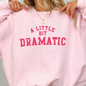 May include: A pink sweatshirt with the text "A Little Bit Dramatic" in red.