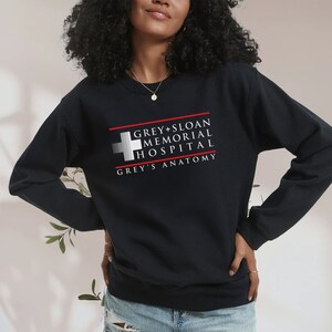 May include: Black sweatshirt featuring the text "GREY + SLOAN MEMORIAL HOSPITAL" and "GREY'S ANATOMY" in white and red. The design includes a white cross symbol. The sweatshirt is a crew neck style.