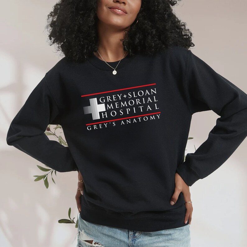 May include: Black sweatshirt featuring the text "GREY + SLOAN MEMORIAL HOSPITAL" and "GREY'S ANATOMY" in white and red. The design includes a white cross symbol. The sweatshirt is a crew neck style.