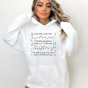 May include: White hooded sweatshirt with a front pocket. The sweatshirt features the text "jingle bells, jingle bells... Probably just gonna jingle some of the way. Oh what comfy it's to hide under a blanket all day" with musical notes and colorful lights.