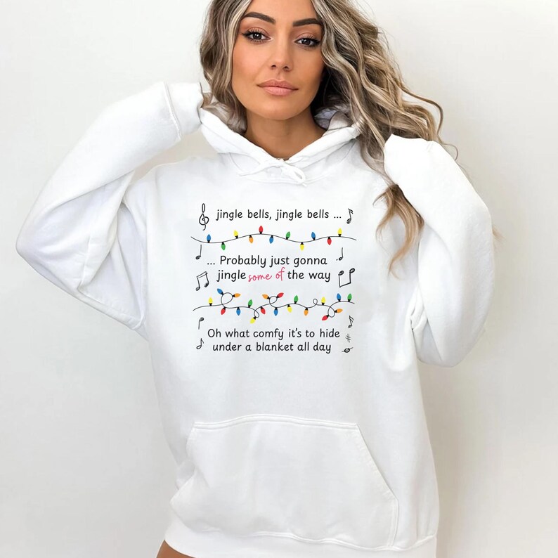 May include: White hooded sweatshirt with a front pocket. The sweatshirt features the text "jingle bells, jingle bells... Probably just gonna jingle some of the way. Oh what comfy it's to hide under a blanket all day" with musical notes and colorful lights.