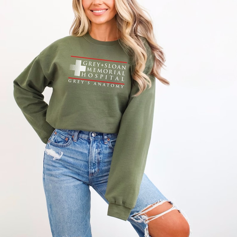 May include: Olive green sweatshirt with the text "GREY+SLOAN MEMORIAL HOSPITAL" and "GREY'S ANATOMY" in white and red. The long-sleeved top is paired with light blue ripped jeans.