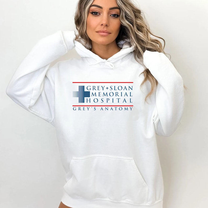 May include: White hooded sweatshirt featuring the text "GREY+SLOAN MEMORIAL HOSPITAL GREY'S ANATOMY" in blue, red, and white. The design includes a blue cross symbol. The sweatshirt has a front pocket and a drawstring hood.