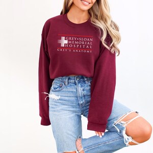 May include: A maroon sweatshirt with the text "GREY+SLOAN MEMORIAL HOSPITAL" and "GREY'S ANATOMY" in white. The sweatshirt has a crew neck and long sleeves. The image shows a person wearing the sweatshirt with blue jeans.