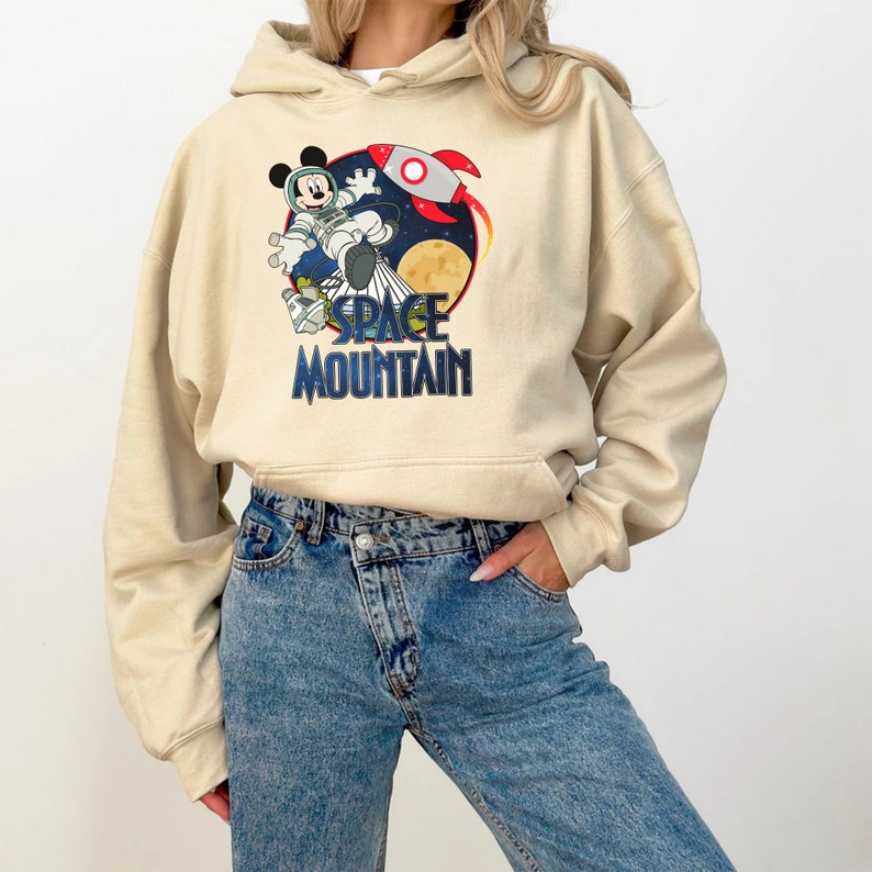 Op de afbeelding: Een beige hoodie met een grafische print van Mickey Mouse verkleed als astronaut met een raket en de tekst "SPACE MOUNTAIN".