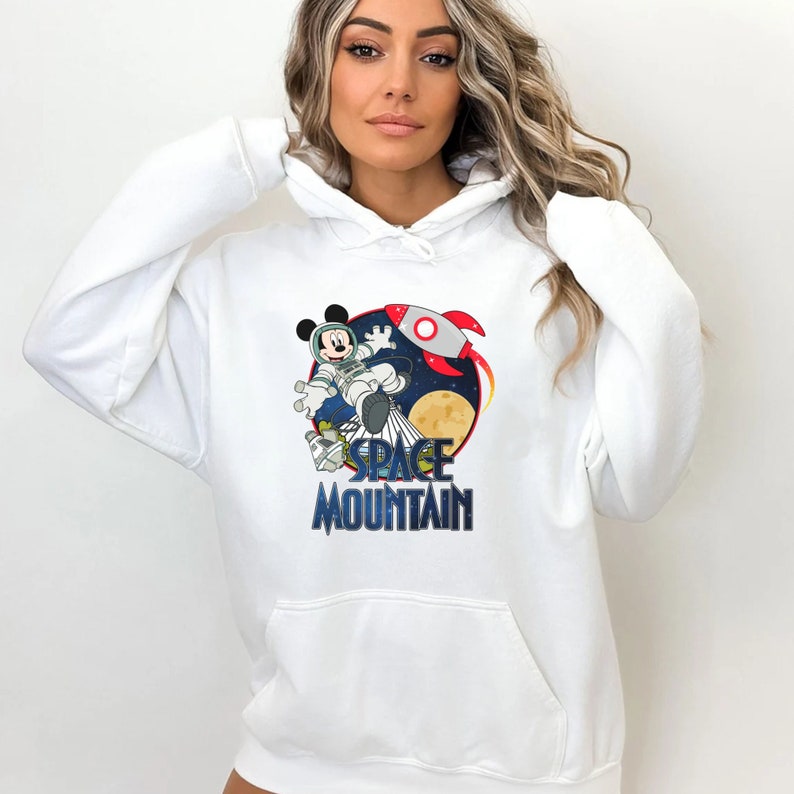 Op de afbeelding: Witte hoodie met een afbeelding van Mickey Mouse als astronaut die door de ruimte vliegt met een raket en de tekst "SPACE MOUNTAIN".