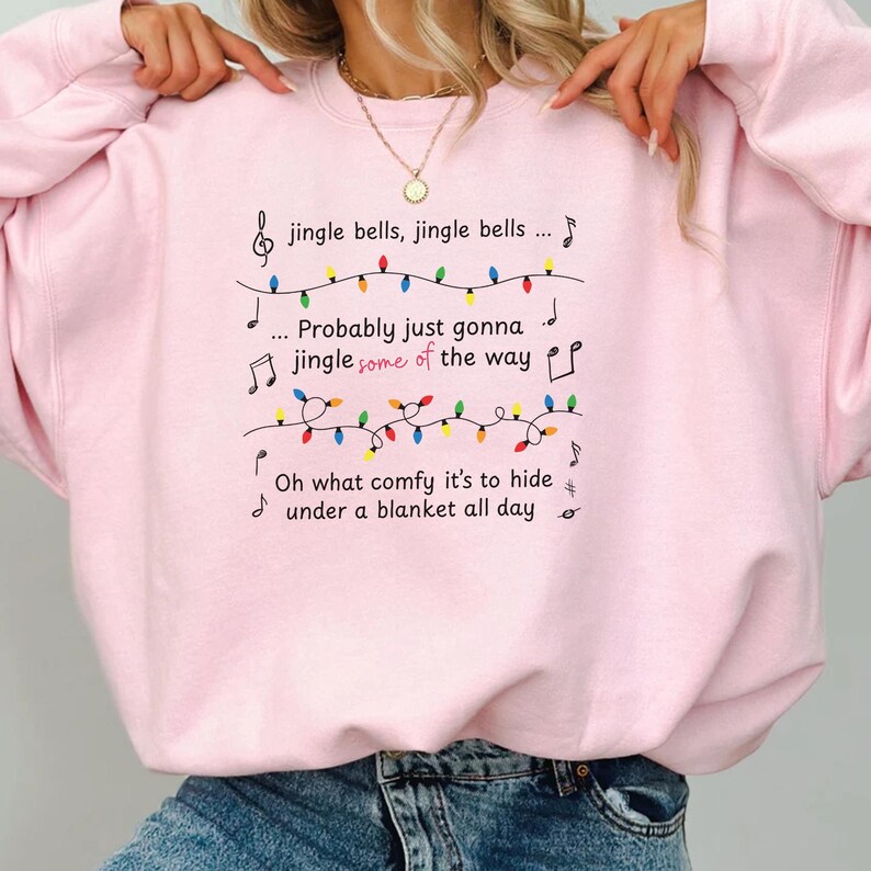 May include: A light pink sweatshirt with black text and musical notes. The text reads "jingle bells, jingle bells... Probably just gonna jingle some of the way... Oh what comfy it's to hide under a blanket all day."