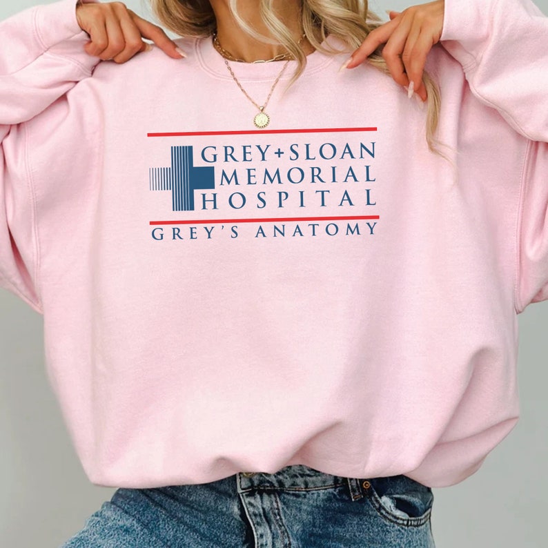 May include: A light pink sweatshirt featuring the text "GREY + SLOAN MEMORIAL HOSPITAL" and "GREY'S ANATOMY" in navy blue and red. The design includes a blue and white cross symbol. The sweatshirt is a crew neck style.