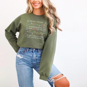 May include: Olive green sweatshirt with the lyrics "Jingle Bells" printed in white. The text includes musical notes and a string of colorful Christmas lights. The sweatshirt is a long-sleeved, crew neck style.