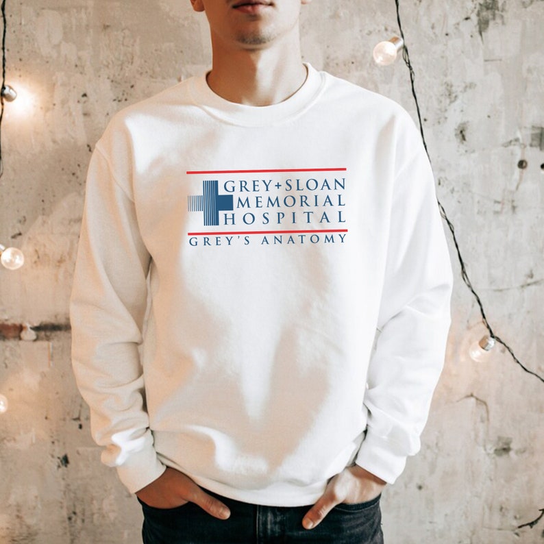 May include: White sweatshirt featuring the text "GREY + SLOAN MEMORIAL HOSPITAL" in blue and red, with a cross symbol. The words "GREY'S ANATOMY" are also included. The sweatshirt is a crew neck style.