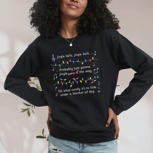May include: Black sweatshirt with white text and musical notes. The text reads "jingle bells, jingle bells... Probably just gonna jingle some of the way... Oh what comfy it's to hide under a blanket all day."