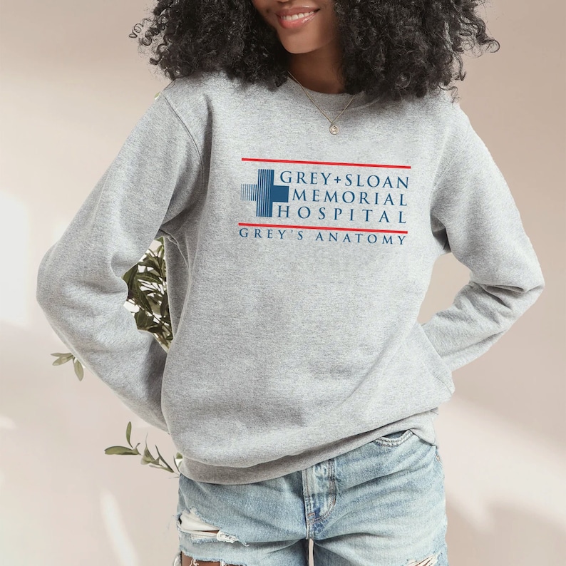 May include: A light gray sweatshirt featuring the text "GREY + SLOAN MEMORIAL HOSPITAL GREY'S ANATOMY" in blue and red. The sweatshirt has long sleeves and a crew neck.