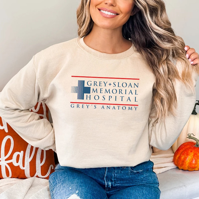 May include: A beige sweatshirt featuring the text "Grey + Sloan Memorial Hospital" and "Grey's Anatomy" in navy blue and red. The design includes a cross symbol. The sweatshirt is a casual style.