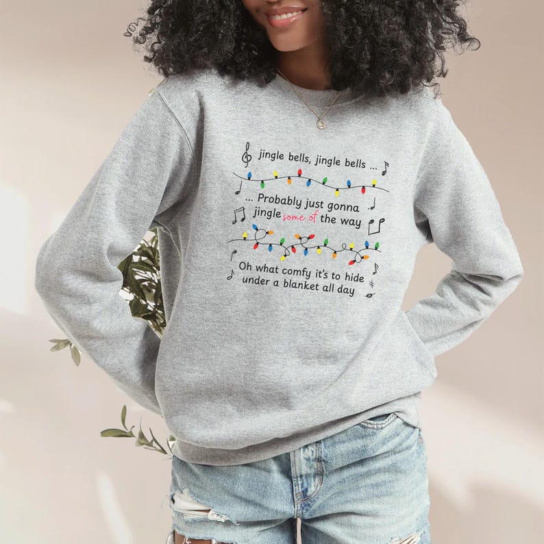 May include: A light gray sweatshirt with the lyrics "jingle bells, jingle bells... Probably just gonna jingle some of the way. Oh what comfy it's to hide under a blanket all day" printed on the front. The text is accompanied by musical notes and a string of colorful Christmas lights.