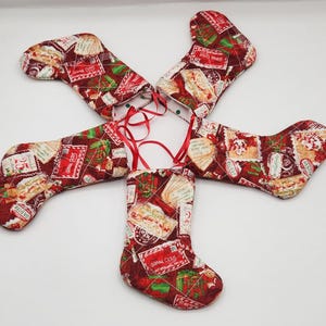 May include: Four fabric Christmas stockings arranged in a circular pattern, each featuring a red background with a repeating print of envelopes, gifts, and text including Santa Claus and Believe. Each stocking has a red ribbon loop for hanging.