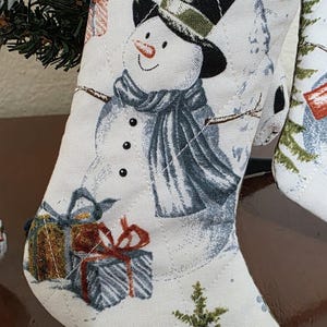 May include: A quilted fabric Christmas stocking in an off-white color featuring a printed snowman wearing a black top hat and a blue scarf. The design includes wrapped gift boxes, a green pine tree, and a small red bird at the bottom edge.