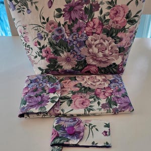 May include: A set of three fabric items featuring a floral pattern with shades of purple, pink, and green on a white background. The set includes a zippered pouch, a rectangular folding case with a purple snap button, and a small key fob pouch.