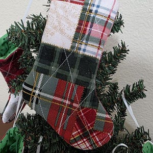 May include: A quilted Christmas stocking featuring a patchwork design of red, green, white, and black plaid fabric patterns. The stocking is constructed with visible white cross-stitch quilting and includes a white ribbon loop for hanging.