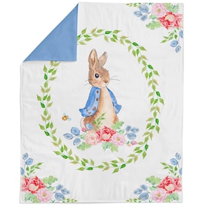 May include: A white blanket featuring a watercolour illustration of a brown rabbit wearing a blue jacket, centred within a green leafy wreath. The design includes pink and red floral accents, blue leaves, and a small bee, with a solid blue corner.