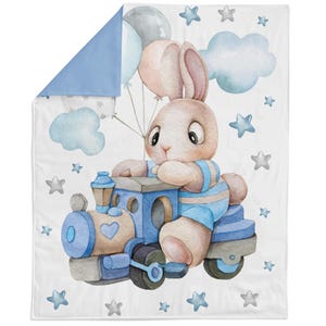 Può includere: Una coperta reversibile per bambini con un'illustrazione ad acquerello di un coniglietto che guida un treno blu e tiene in mano dei palloncini. La coperta ha un lato bianco con stelle e nuvole e un lato reversibile blu chiaro.
