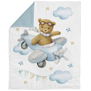 May include: A baby blanket featuring a watercolor illustration of a teddy bear wearing goggles and a scarf, flying a light blue airplane. The design includes clouds, stars, and a banner on a white background with a light blue border.