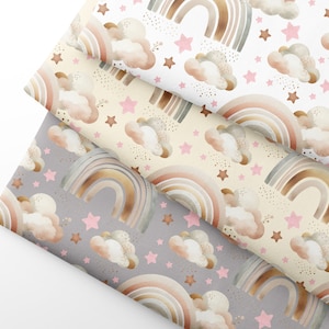 Può includere: Tre campioni di tessuto con un motivo ripetuto di arcobaleni ad acquerello, nuvole e stelle. Gli arcobaleni sono in tonalità di marrone, rosa e verde. Le nuvole sono rosa chiaro e le stelle sono dorate e rosa. I campioni sono su uno sfondo bianco.