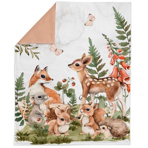 Op de afbeelding: Een rechthoekige deken met een aquarelillustratie van een reekalf, vos, konijn, muis en egel in een bosrijke omgeving met groene varens, rode vingerhoedskruidbloemen, aardbeien en vlinders op een witte achtergrond met een perzikkleurige hoek.