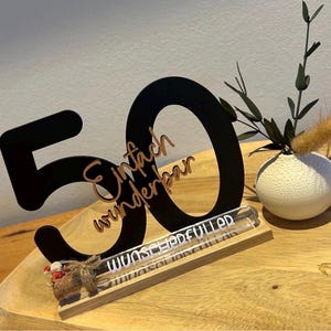 May include: A black wooden "50" centerpiece with the words "Einfach wunderbar" in a cursive font. A clear glass tube with text sits on a wooden base. A small white vase with greenery is to the right.