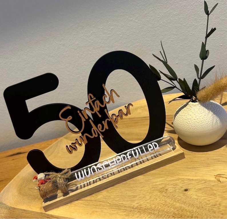 May include: A black wooden "50" centerpiece with the words "Einfach wunderbar" in a cursive font. A clear glass tube with text sits on a wooden base. A small white vase with greenery is to the right.