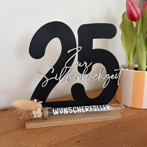 May include: A black wooden '25' centerpiece with the words 'Zur Silberhochzeit' in silver script. A small glass tube with the word 'WUNSCHERFUELLER' sits on a wooden base, with dried flowers.
